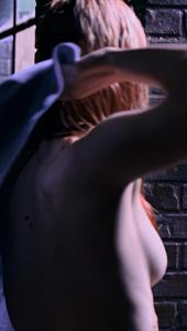 A moody, dimly lit shot from Daredevil Season 1, Episode 1. Deborah Ann Woll is caught mid-motion with her arms raised, creating a natural, unposed angle that reveals a subtle glimpse of sideboob.